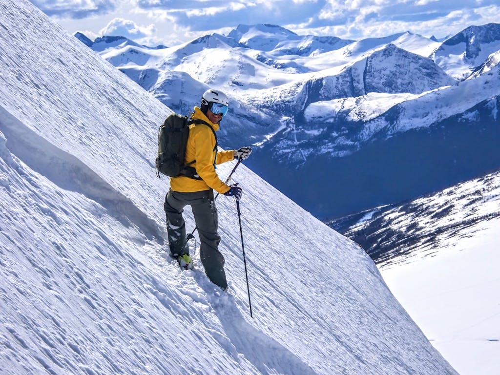 A skier in a yellow jacket descending a steep snowy slope with mountains in the background.