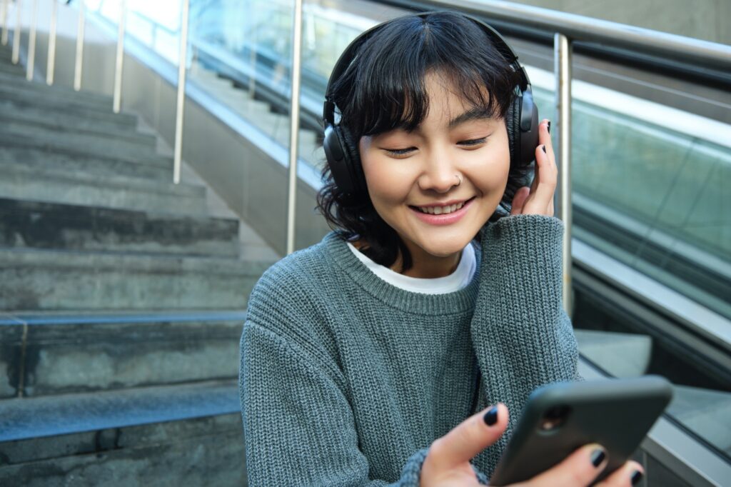 Portrait of smiling korean girl in headphones, uses smartphone and sits on stairs in mall, watches