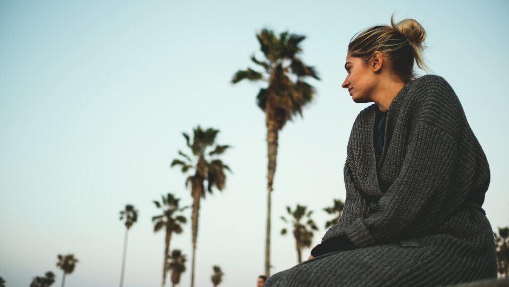 About 1 woman wearing gray sweater sitting with the distance of coconut trees under white cloudy sky